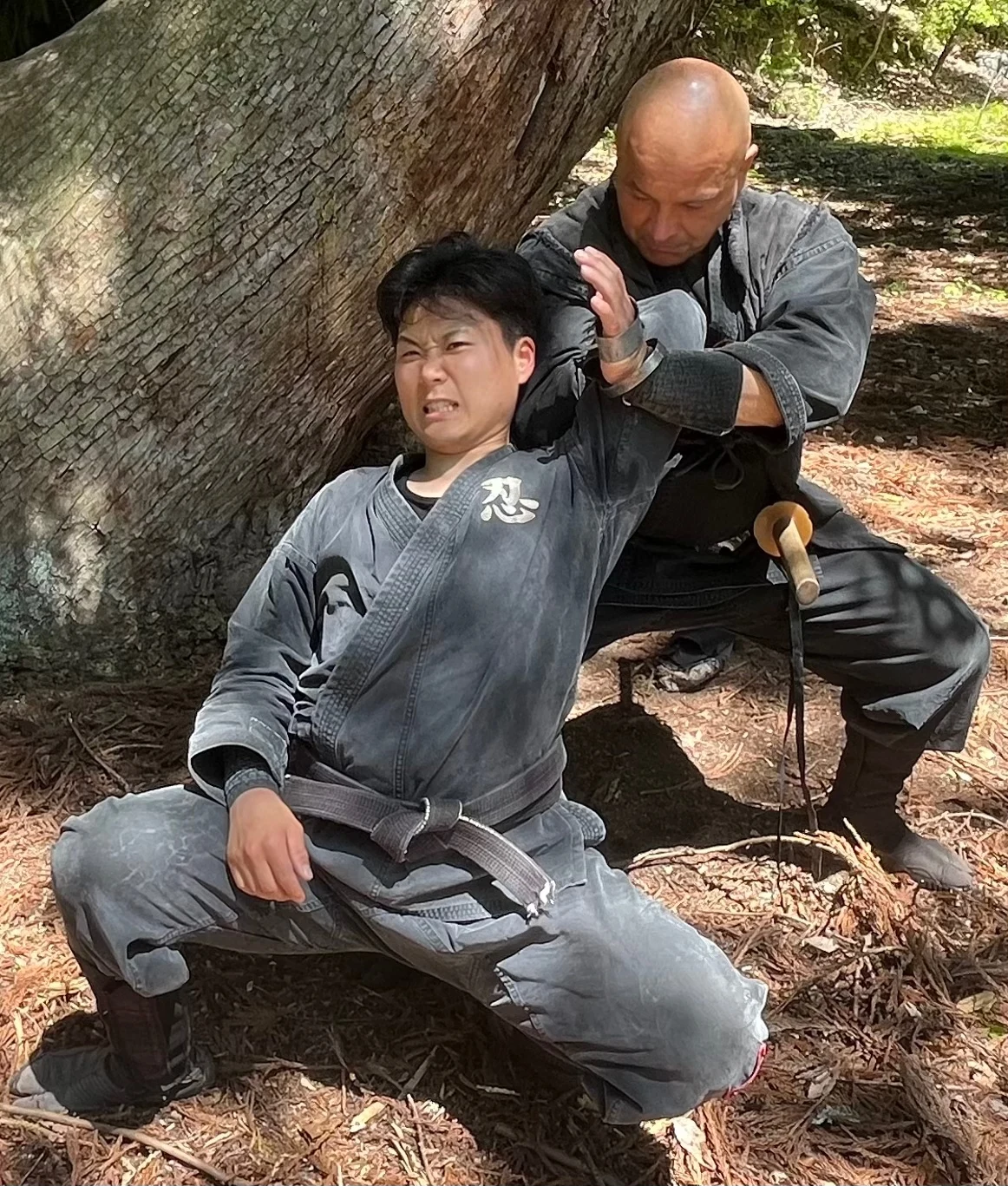 木の前に座る稽古者と、その後ろから頭部の近くへ手を添える相手。